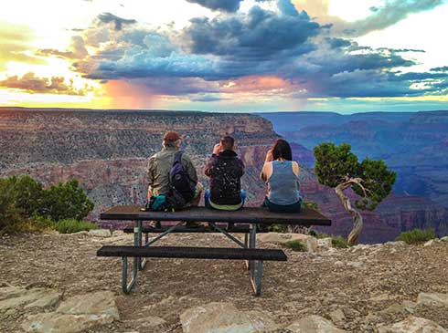 Three tourists seated atop picnic table watching sweeping rainbow colored sunset over Grand Canyon South Rim