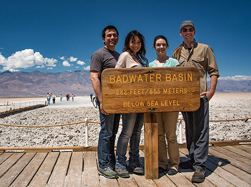 Four people posing by Badwater Basin elevation sign at Death Valley National Park with salt pans in background.