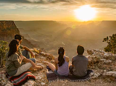 Family of four seated at the edge of the Grand Canyon's South Rim watching a golden sunset.