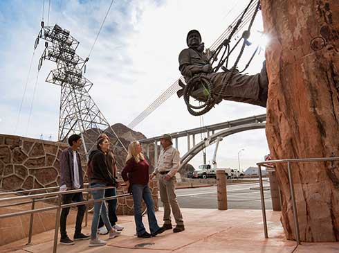 Group of Pink Jeep tour guests with guide looking at the bronze High Scaler Monument at Hoover Dam