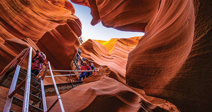 Tour visitors descending the steep metal staircase into Lower Antelope Canyon, with crack of blue sky at top.