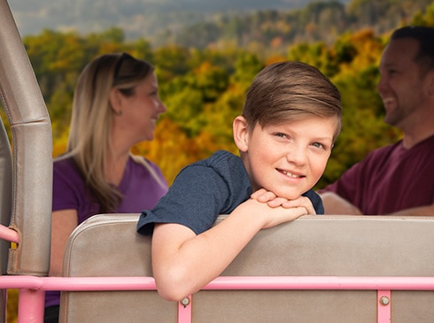 Young boy looking at the camera from the back of a Pink Jeep Wrangler, with parents and fall-colored leaves in the background.
