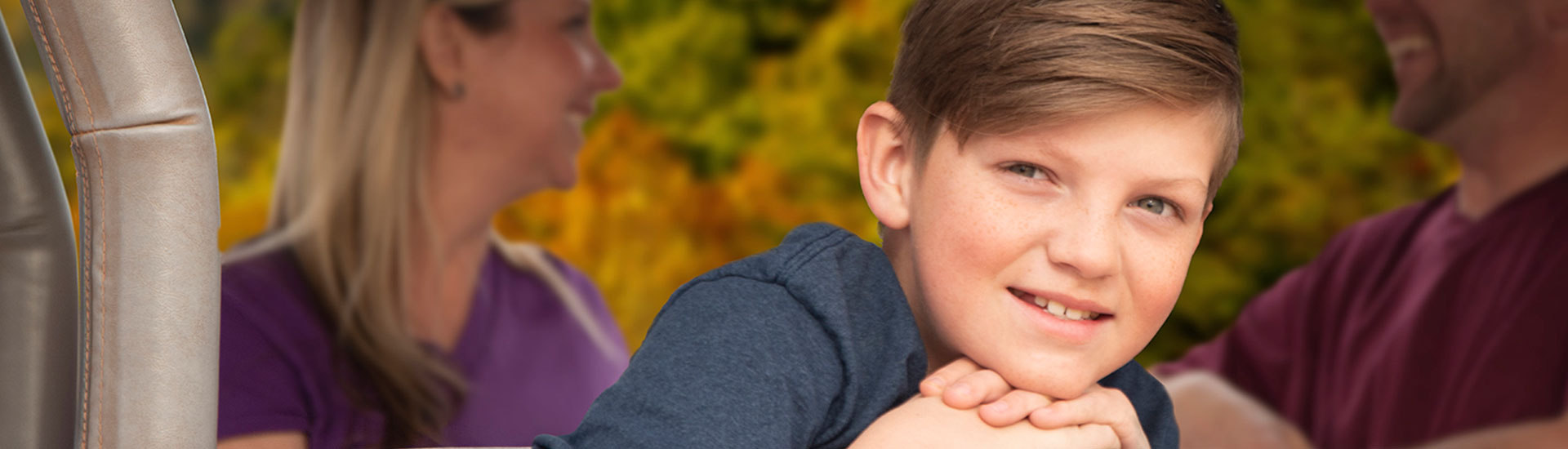 Close-up of young boy looking at camera from the back of a Pink Jeep Wrangler, with parents and fall-colored leaves in background.