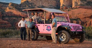 Tour guide and guests beside a Pink Jeep Wrangler, looking golden sunlight falling on red rocks in background.