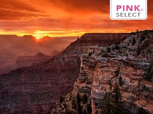 Vibrant orange sunset at South Rim of Grand Canyon National Park with rock cliffs in foreground and Pink Select logo.