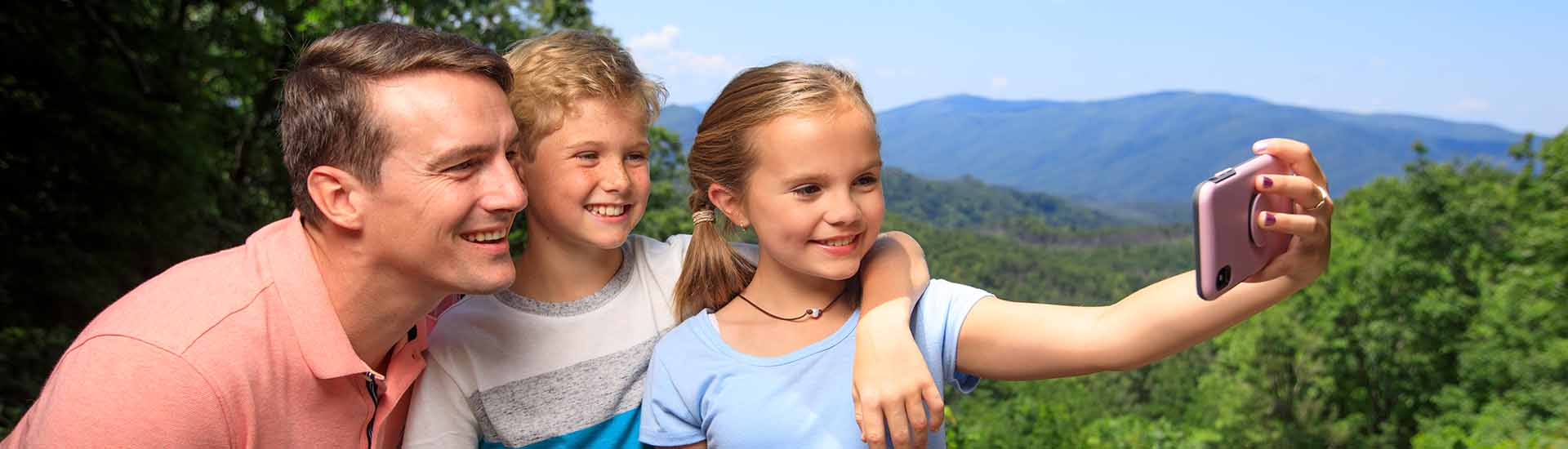 Father, son and daughter taking selfie with Smoky Mountains' hilltops and blue sky in background on beautiful summer day