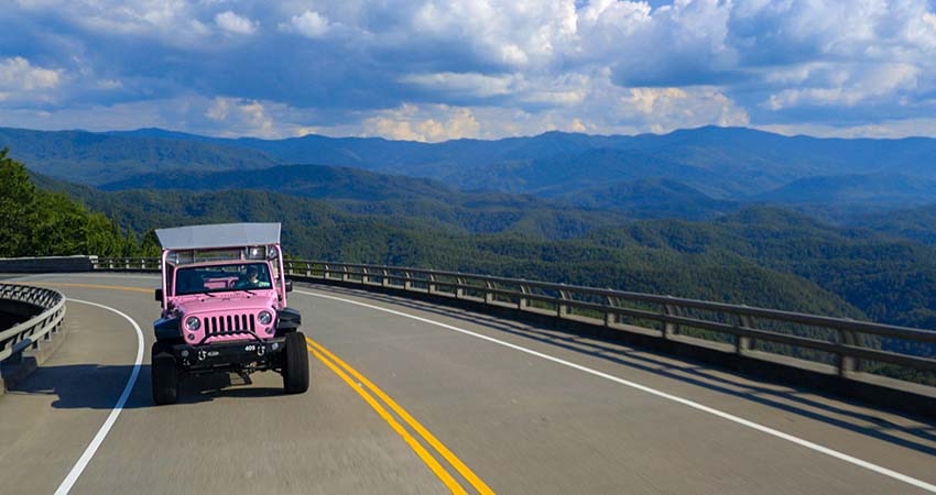 Pink Jeep traveling towards camera on Foothills Parkway with Smoky Mountains and puffy clouds in background