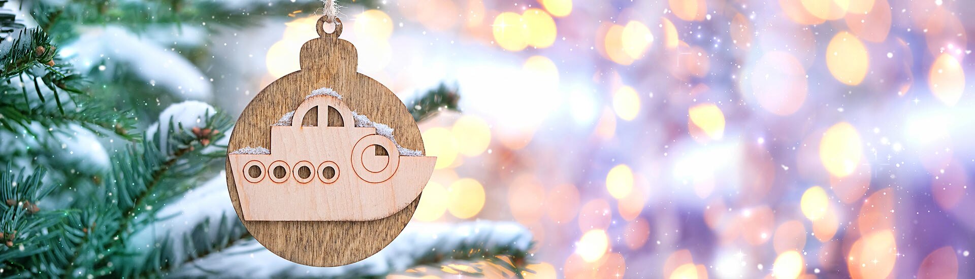 Wooden medallion ornament with carved boat hanging on snowy Christmas tree limb, yellow-pink lights glow in background.