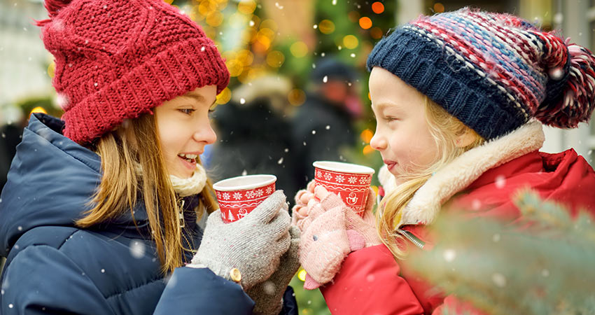 Two adorable sisters drinking hot chocolate with snow falling on a traditional Christmas plaza in Smoky Mountains.