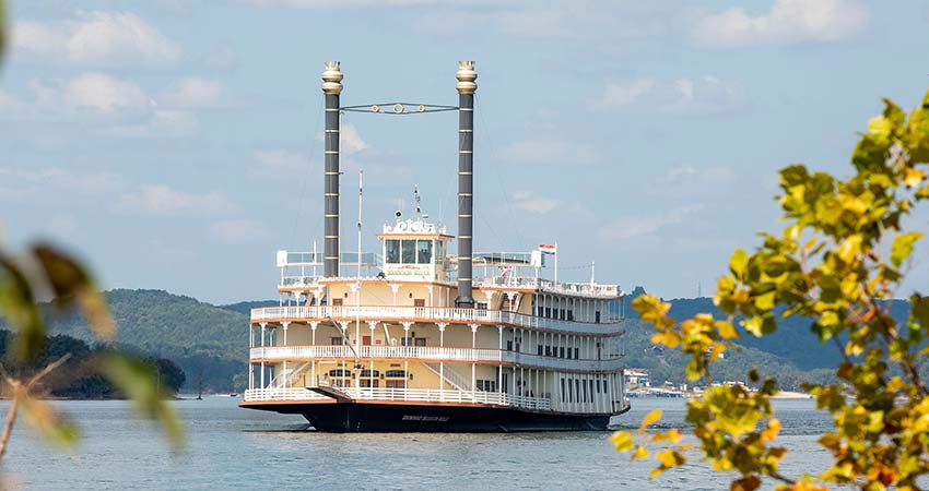 The Branson Belle Showboat floating on the blue waters of Table Rock Lake, framed by trees in the foreground.