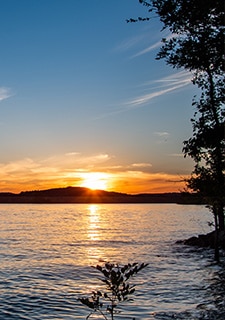 Golden sun sets below the Branson hills, with water ripples of Table Rock Lake and shoreline trees in the foreground.
