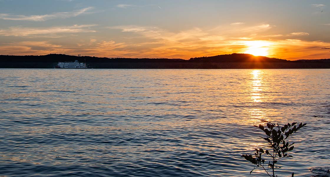 A golden sun dips below the Branson shoreline, reflecting across Table Rock Lake with the Branson Belle in the distance.