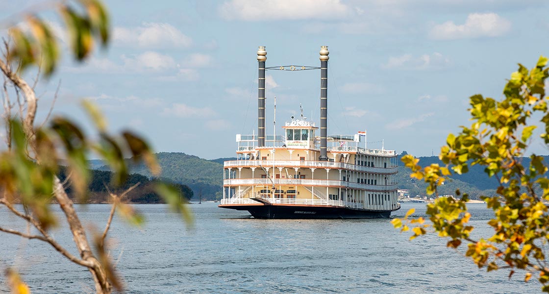 Showboat Branson Belle floating on the waters of Table Rock Lake seen through shoreline tree branches in the foreground.