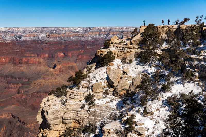 Tiny people stand atop the edge of the Grand Canyon's South Rim, its sandstone cliffs lightly dusted in snow.