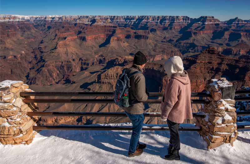 A man and woman gaze at the rust-colored walls of the Grand Canyon with a blanket of snow beneath their feet.