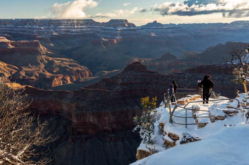 A family standing on a snow-covered Grand Canyon viewpoint in morning light, looking into the depths of nearby canyon walls.
