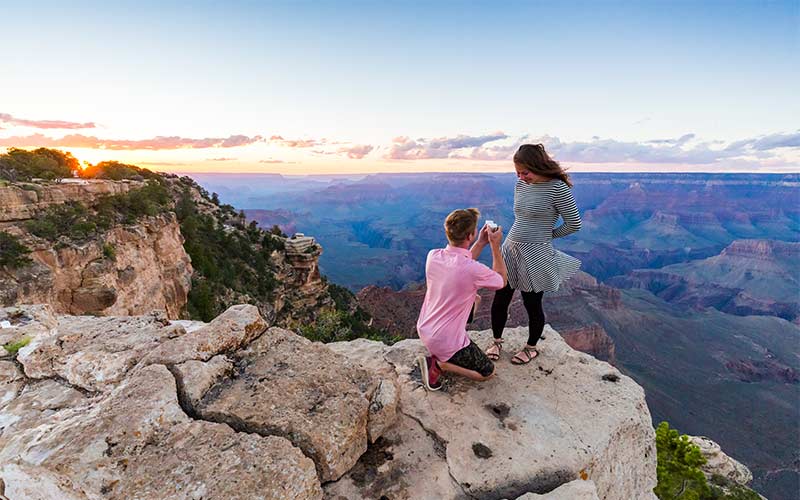 Young man proposing to his girlfriend on a rock outcropping overlooking the Grand Canyon at sunset.