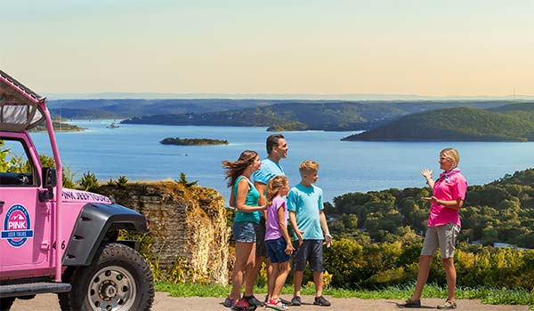 Tour guide and guests at a scenic viewpoint atop Baird Mountain overlooking Table Rock Lake, with Pink Jeep Wrangler in the foreground.