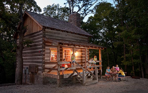 A family gathers around the softly lit porch of a rustic log cabin at Silver Dollar City Campground at dusk.