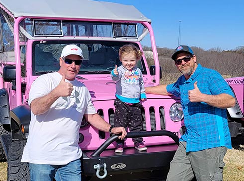 A toddler stands on the bumper grill of a Pink Jeep Wrangler with two males standing alongside giving a thumbs-up sign.