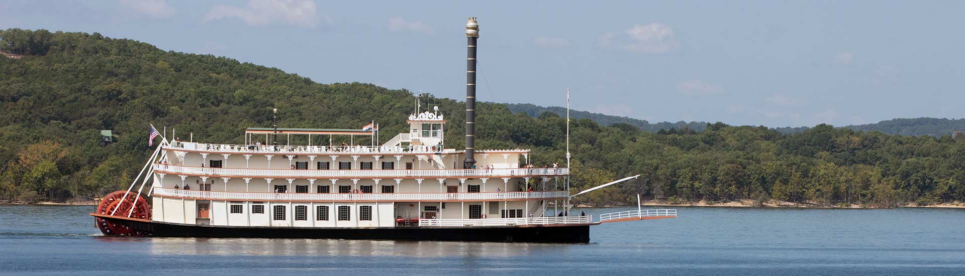 The Branson Belle Showboat glides across Table Rock Lake with its paddle wheel in motion and the Branson hills in background.
