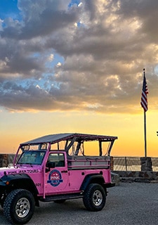 A Pink Jeep Wrangler is parked near a flagpole atop Baird Mountain as a golden sunset casts shadows on the clouds above.