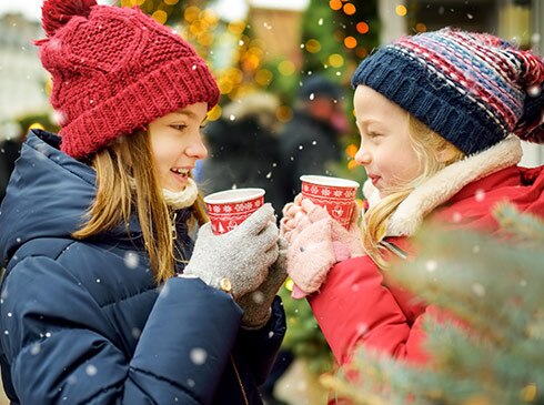 Two young girls bundled up in red and blue jackets among falling snowflakes, drinking warm wassail in a Christmas square.