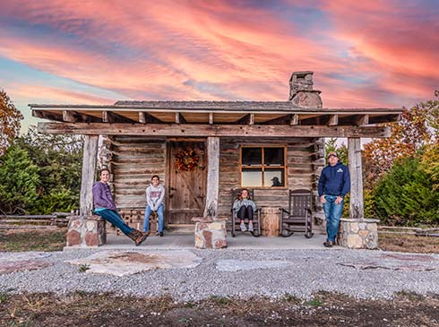 Family of four poses on the porch of a hand-hewn log cabin during a pink sunset while on a Pink Jeep Tour, Branson, MO.