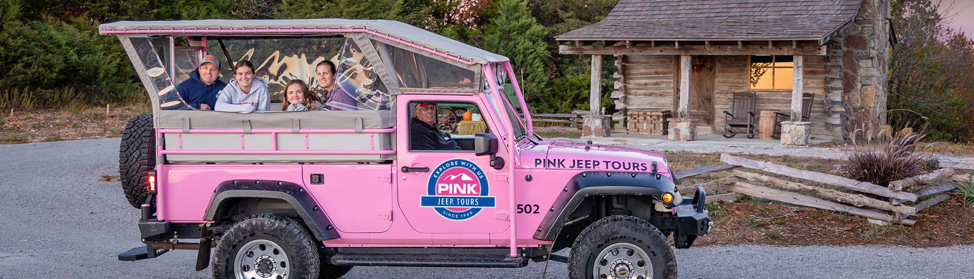 Tour guests smiling from the back of a Pink Jeep Wrangler parked in front of historic log cabin at sunset, Pink Jeep Tours Branson.