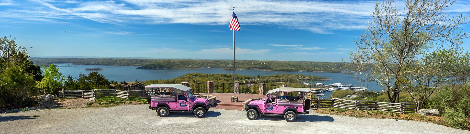 Two iconic Pink Jeep Wrangler parked by flagpole at Pink Jeep Tours’ Baird Mountain lookout with Table Rock Lake in background.