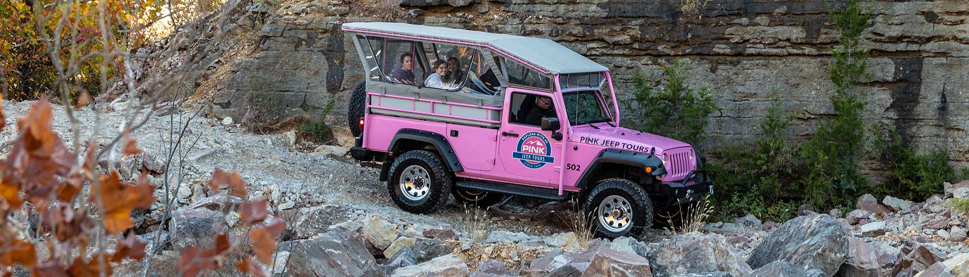 Side view of a Pink Jeep Wrangler traveling down a rocky trail along the stone face of Baird Mountain, with autumn trees in foreground.
