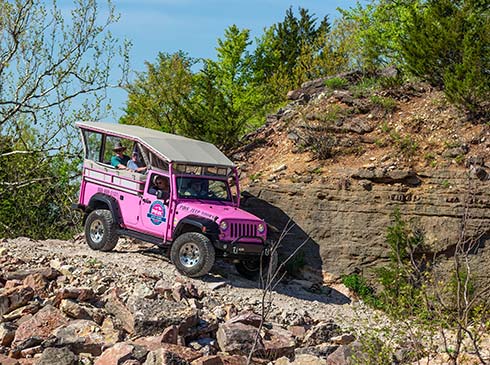 A Pink Jeep Wrangler rounds a cliff side corner of Baird Mountain along a rocky 4x4 trail, with trees and blue sky in background.
