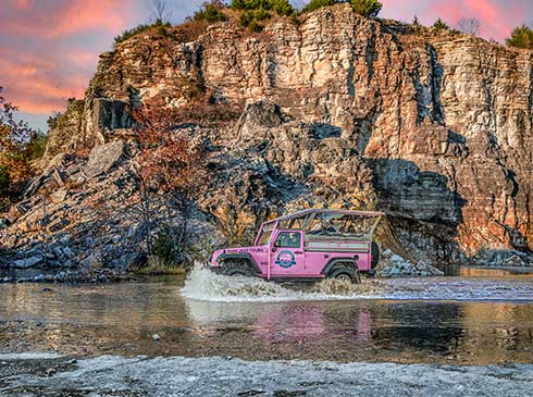 A Pink Jeep Wrangler crosses still waters in front of the Baird Mountain rockface during a pink sunset, Pink Jeep Tours Branson.