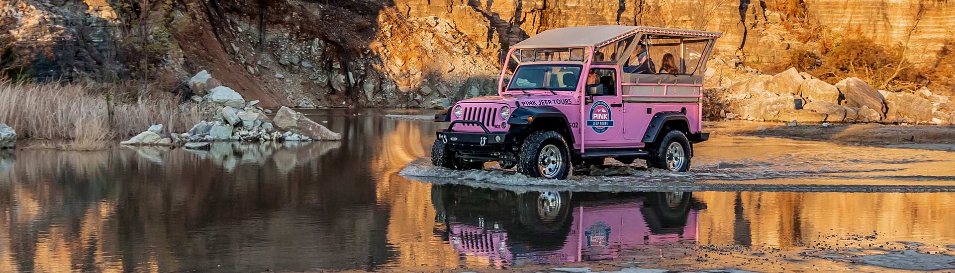 Pink Jeep Tours’ Jeep Wrangler glides through still water in front of a golden-lit rockface with its reflection below.