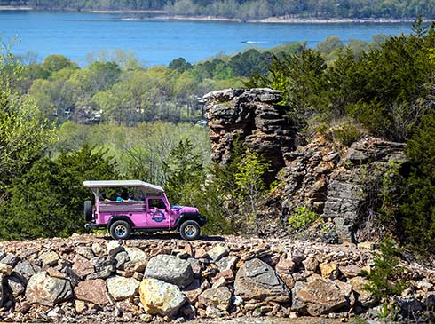 A Pink Jeep Wrangler traverses a 4x4 trail near tall rocky outcrops with a green forest and Table Rock Lake in the background.