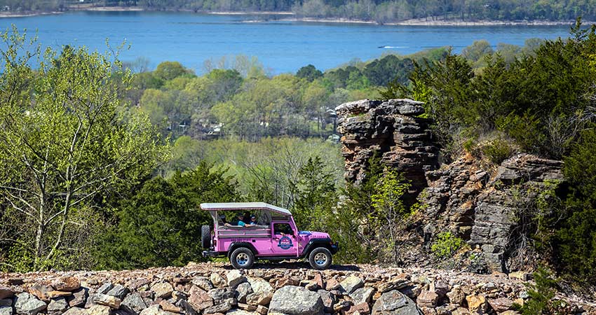 A Pink Jeep Wrangler on a 4x4 trail approaches two tall rock outcrops framed by forested trees and Branson’s Table Rock Lake.