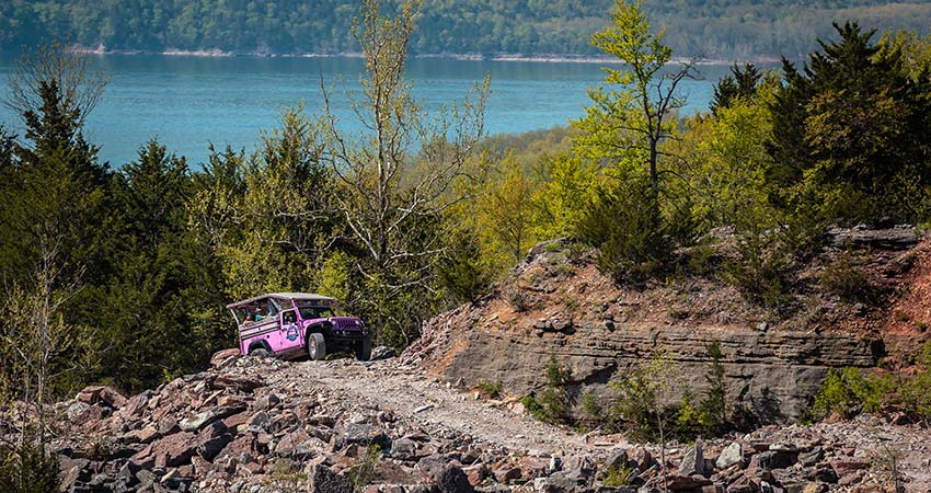 A Pink Jeep Wrangler climbs a 4x4 trail in the rock quarry below Baird Mountain with forested trees and Table Rock Lake in the background.