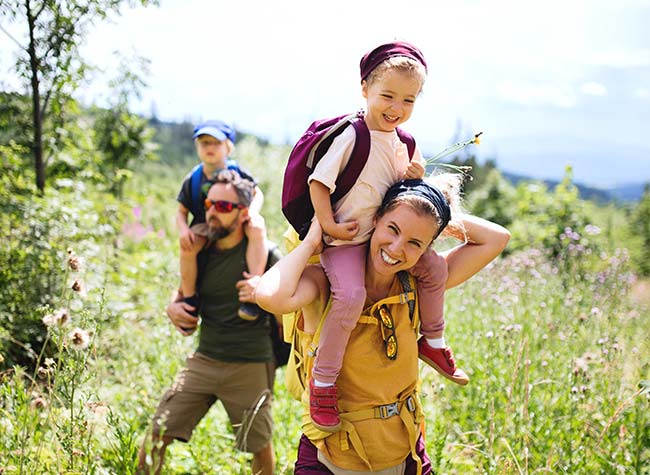 Family hiking through the hills of a blooming meadow with forest trees and mountains in background.