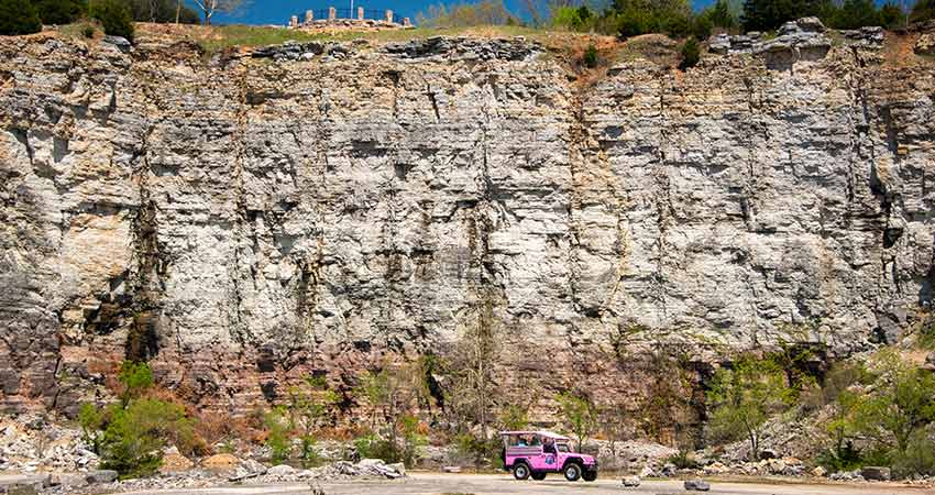 Pink Jeep Wrangler at bottom of Ozark mountain quarry, with tall cliff face of Baird Mountain in background.