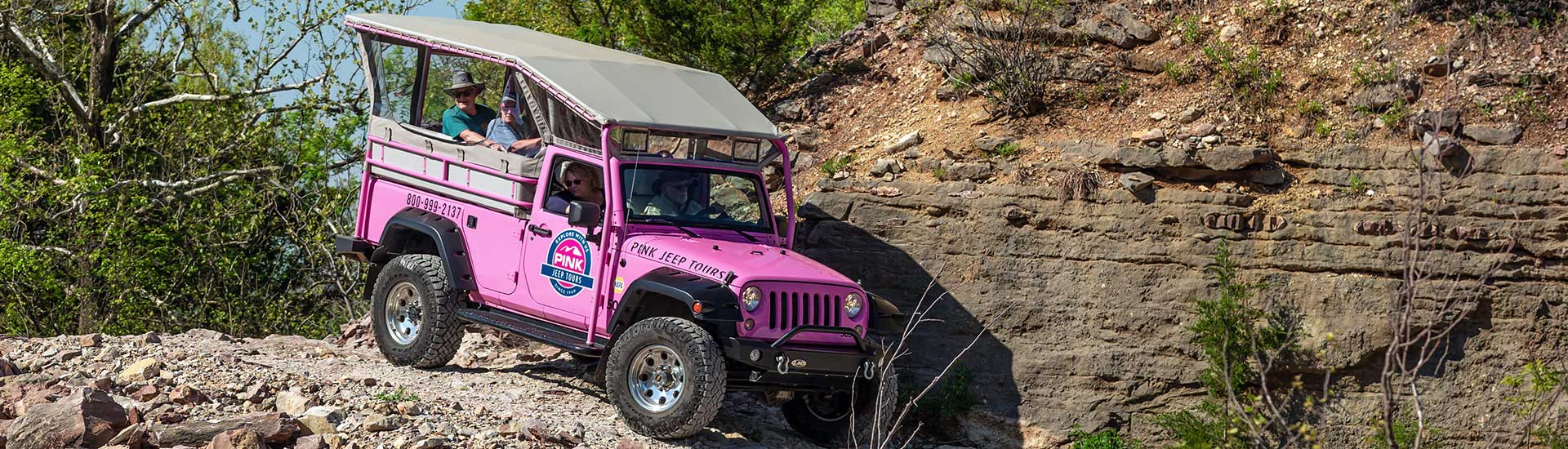 Side view of tour guests inside a Pink Jeep Wrangler as it winds along the rocky trail beside cliff face of Baird Mountain.