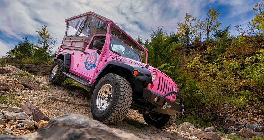 Closeup side frontal view of Pink Jeep Wrangler perched on a steep rocky trail, blue sky in background,