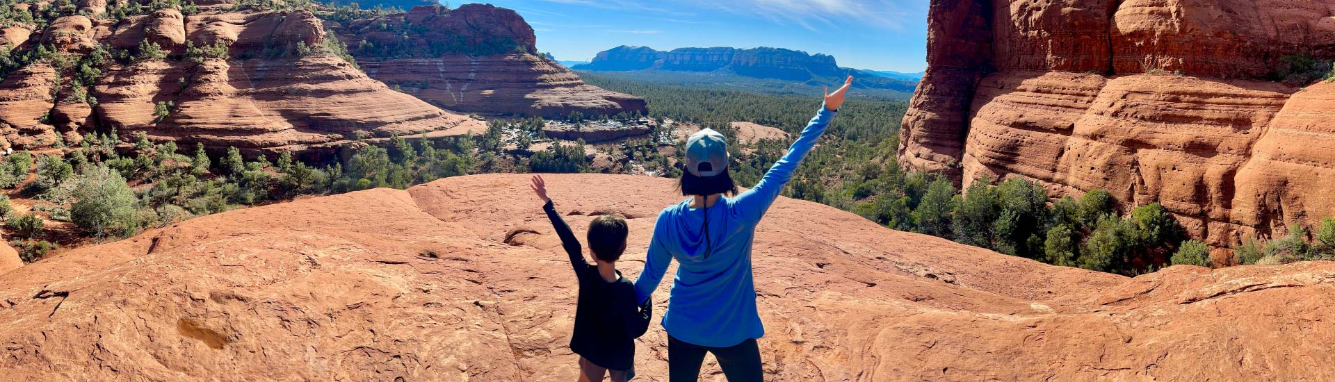 Mother and son stand on a rock looking out over Broken Arrow Trail, arms raised in celebration