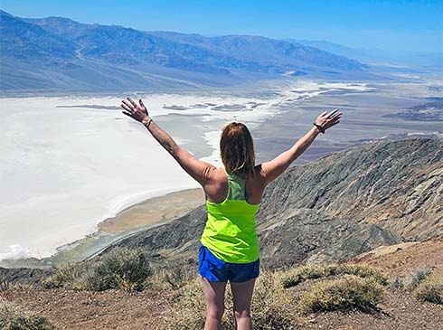 Woman standing with arms spread wide as she looks down on the colorful salt pans of Death Valley National Park from Dante’s View.