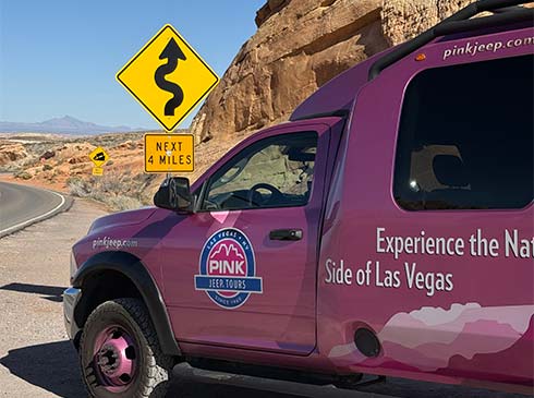 Pink Jeep Las Vegas Tour Trekker parked by a winding road sign in the Valley of Fire State Park, with rocky formations and distant mountains under blue sky.