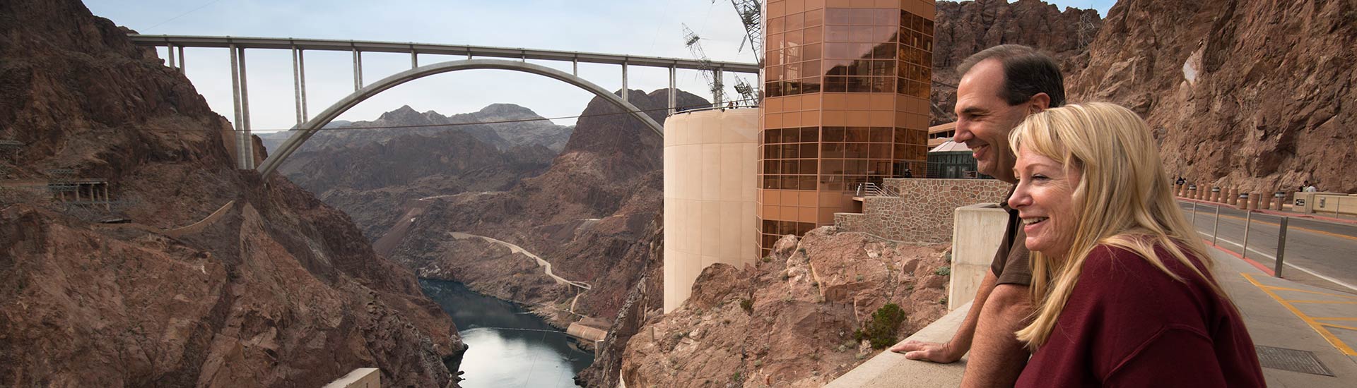 Two tourists on the walkway at Hoover Dam looking at the Colorado River below and the Mike O'Callaghan-Pat Tillman Bridge.