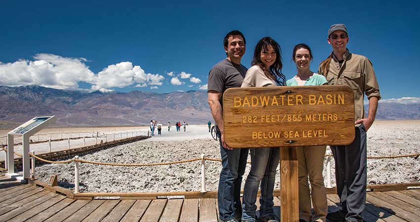 Four people posing behind the Badwater Basin elevation sign with the Death Vally salt flats and mountains in background.