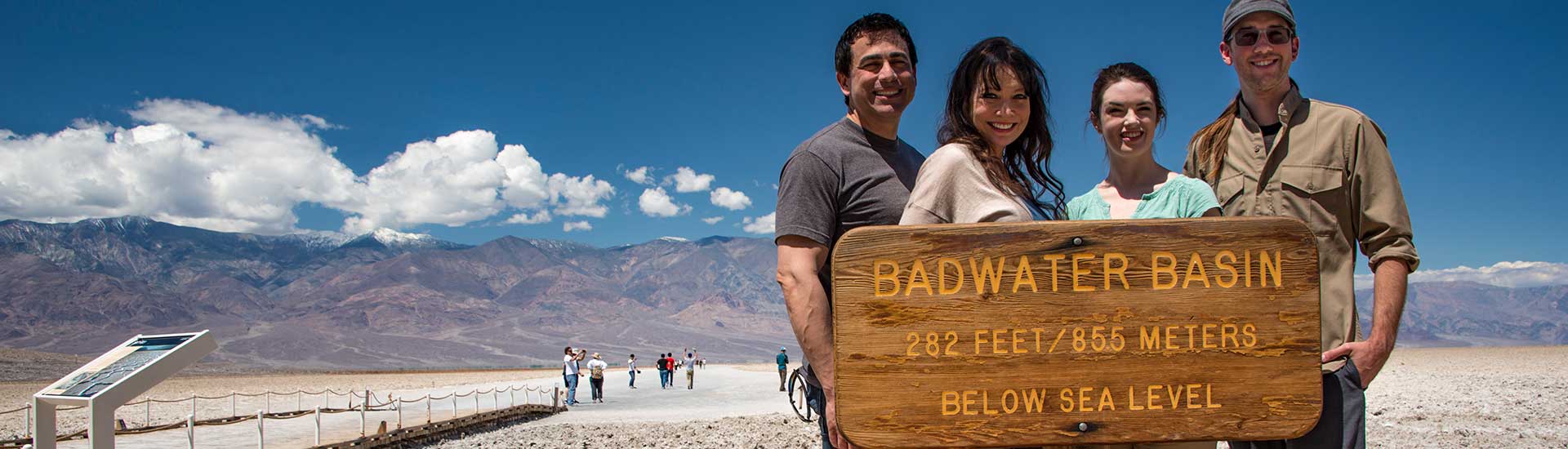 Four people posing by the Badwater Basin elevation sign at Death Valley National Park, with salt flats in background.