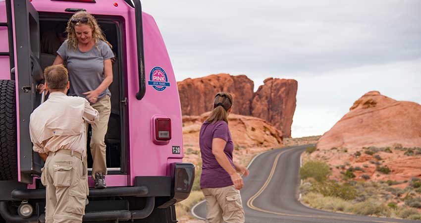 Two women exiting the back of a Pink Jeep Tour Trekker with the guide's help at a tour stop in Valley of Fire State Park, Nevada.