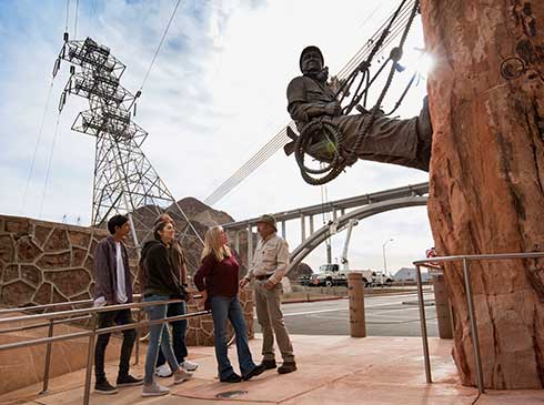 Group of Pink Jeep tour guests with guide looking at the bronze High Scaler Monument at Hoover Dam