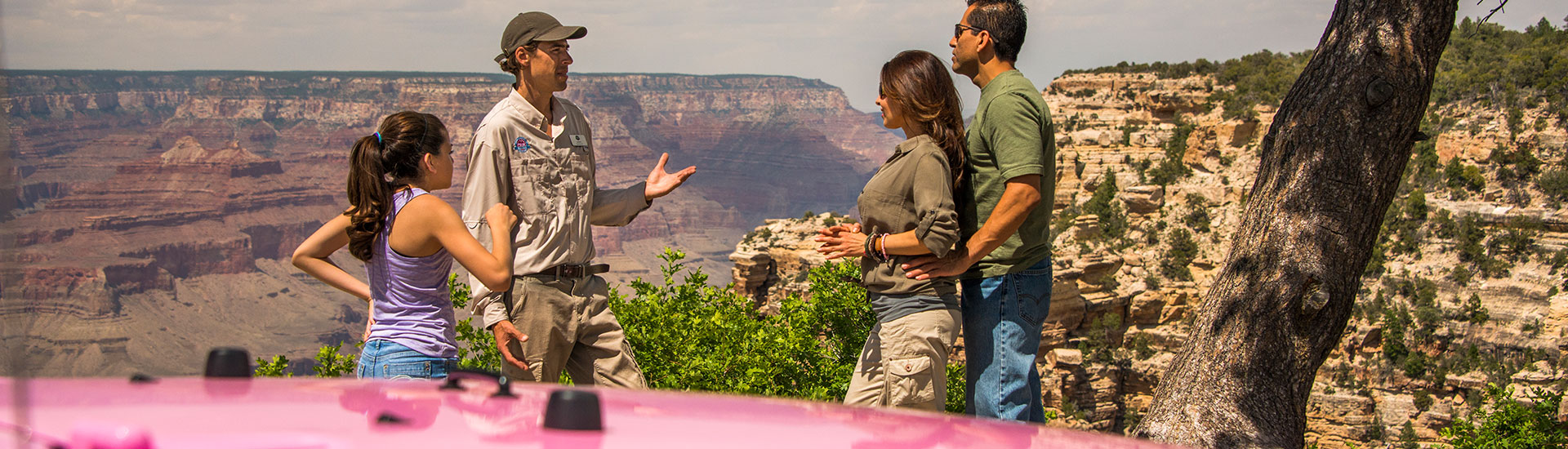 PInk Jeep Tours' Grand Canyon adventure guide talks with a family while standing at a South Rim viewpoint, with the hood of a pink Jeep Wrangler in foreground.